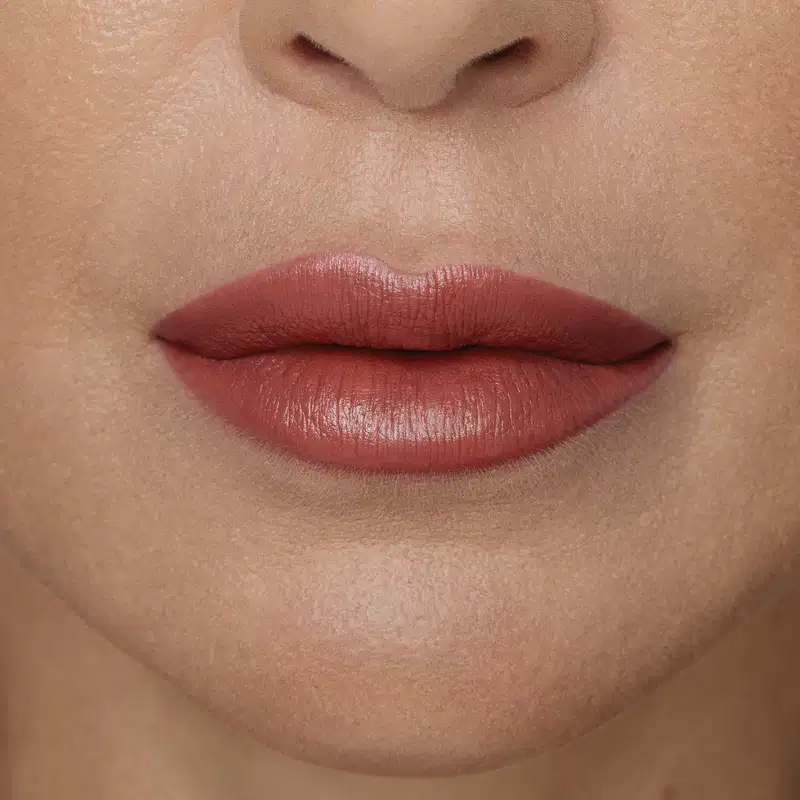 Boca feminina de mulher branca em close mostrando queixo e um pedaço do nariz, com boca fechada, expressão séria e lábios com batom rosado sobre eles.