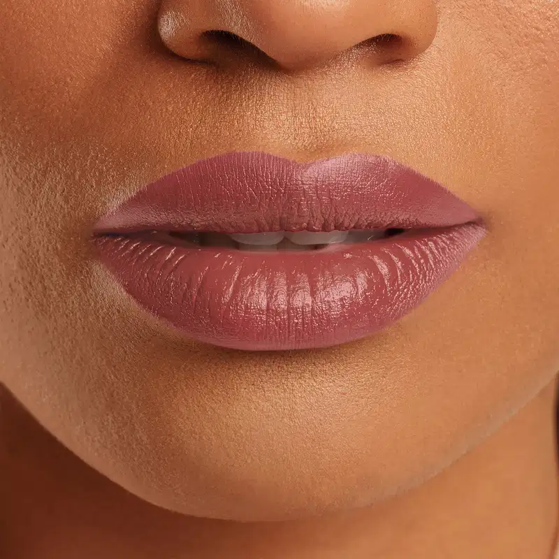 Boca feminina de mulher branca em close mostrando queixo e um pedaço do nariz, com lábios levemente entreaberto, revelando um pouco dos dentes frontais brancos e lábios com batom rosado sobre eles.
