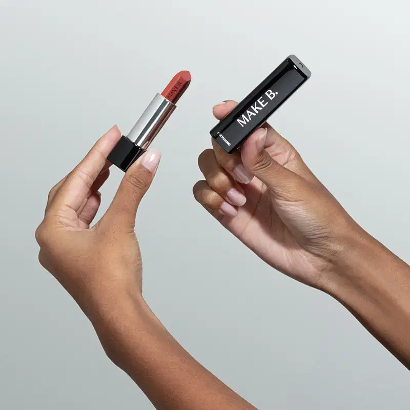 Mãos femininas segurando a tampa preta de um batom da marca Make B. na mão direita e a base do batom com a bala aparente na mão esquerda, em um ambiente minimalista e claro.