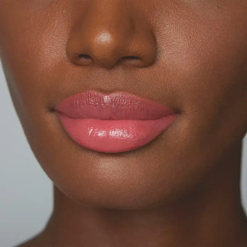 A imagem mostra parte de um rosto feminino em primeiro plano, dando destaque aos lábios, mostrando como o batom rosa da /Make B. fica na pele negra. O fundo é cinza e liso.
