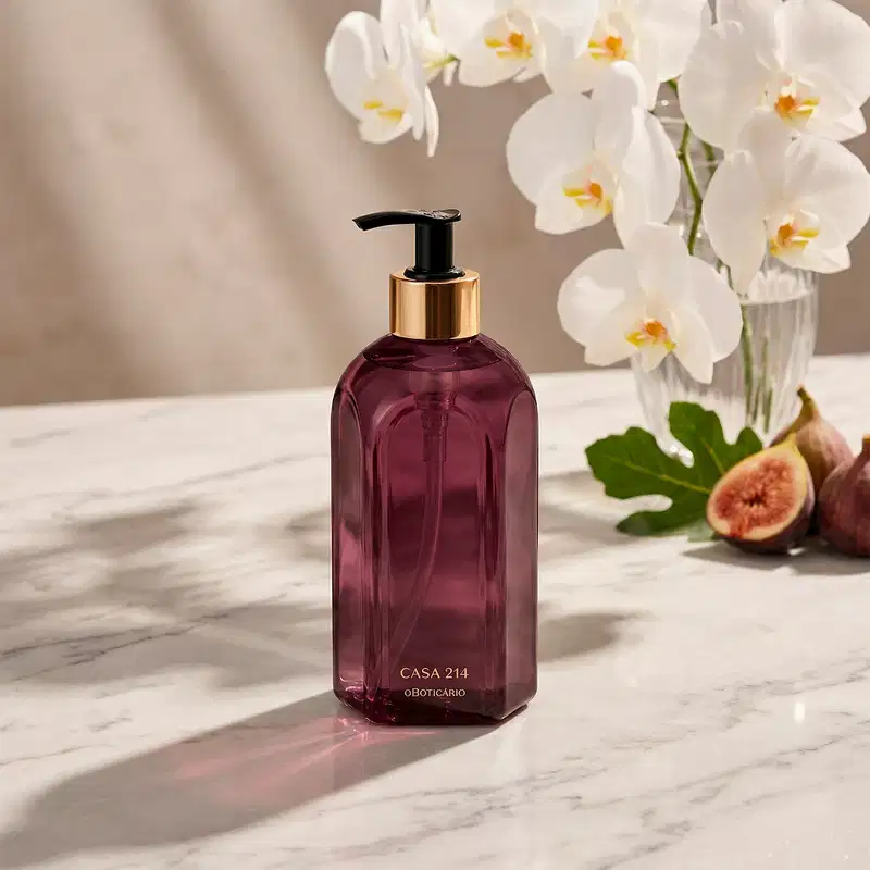 A imagem mostra um vidro roxo do sabonete líquido Casa 214 Figo Magnífico, com bomba dosadora preta. Ele está em superfície de mármore branco, onde o produto faz uma sombra. Existem flores brancas e figo ao fundo, em ambiente iluminado e harmonioso.