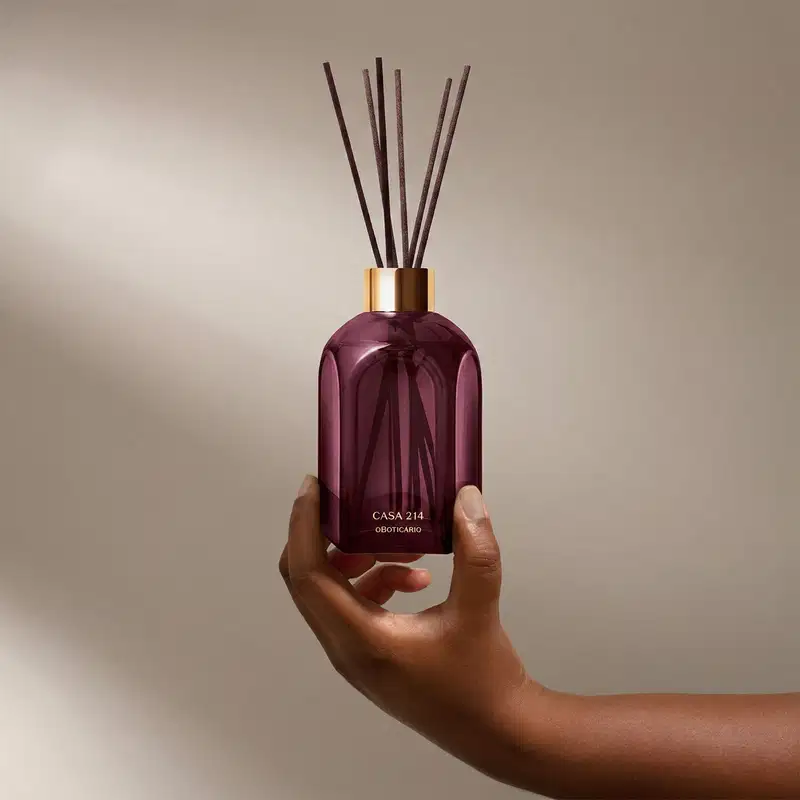 Frasco de difusor de aromas da marca Casa 214, com design geométrico em vidro de cor roxa escura e tampa dourada. Contém varetas de difusão pretas inseridas no frasco.