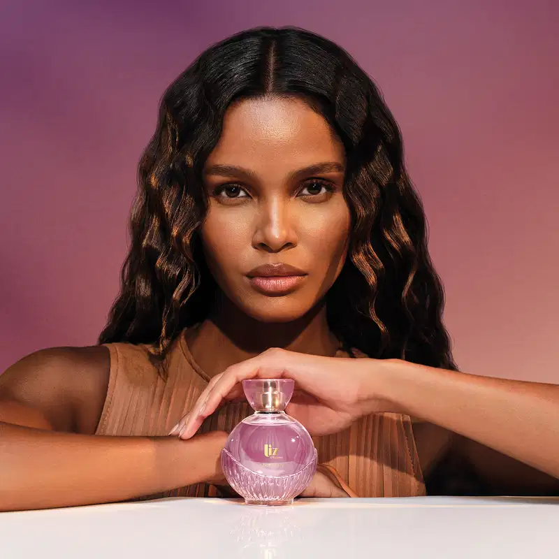 Modelo negra com cabelos longos e ondulados e expressão séria vista dos ombros para cima. A frente dela, um frasco de desodorante colônia da marca Liz D'Essence, sobre o qual ela repousa sua mão esquerda.