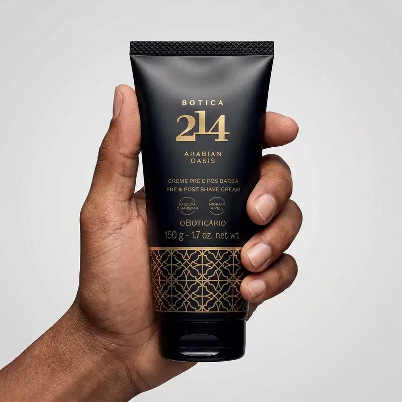 Tubo preto com texto e arabescos dourados da marca Botica 214 Arabian Oasis em fundo branco, sendo segurado por mão masculina de pele parda.