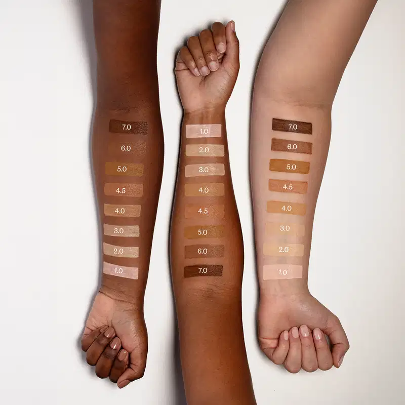 Três antebraços com diferentes tons de pele exibem faixas de diferentes cores de protetor solar e seus respectivos números, sugerindo uma paleta de tons de pele.