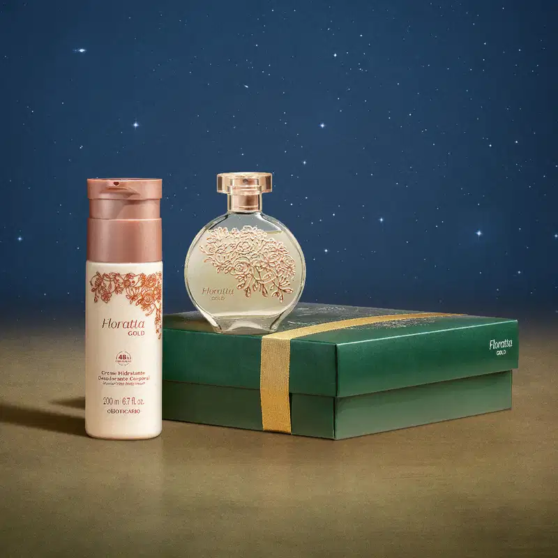 Frasco de vidro de desodorante colônia Floratta Gold sobre  caixa presenteável verde com ilustração natalina. Ao lado um frasco branco com tampa dourada de body splash da mesma linha sobre superfície marrom e fundo azul-escuro simulando um céu estrelado.