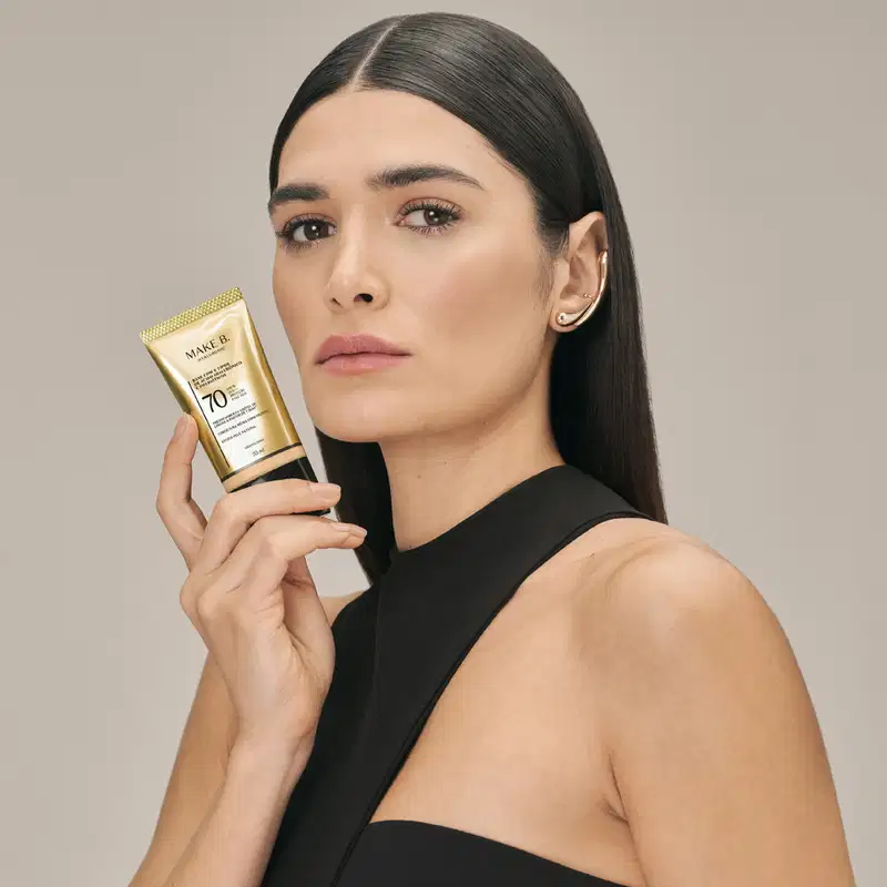 A modelo segura um tubo dourado de base líquida com protetor solar da marca Make B., em um ambiente minimalista e neutro, destacando o produto de beleza.