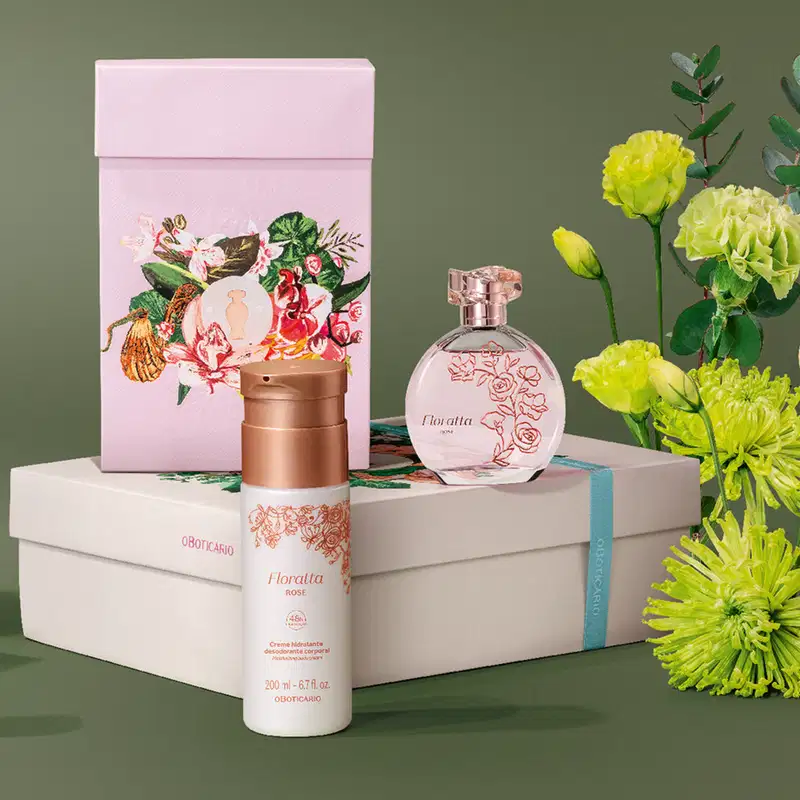 Imagem do kit Floratta Rose, com o clássico vidro transparente do desodorante colônia e frasco branco creme hidratante corporal, além de uma caixa com estampa floral. As embalagens trazem flores e tampas douradas. Ao lado, flores amarelas e fundo cinza.