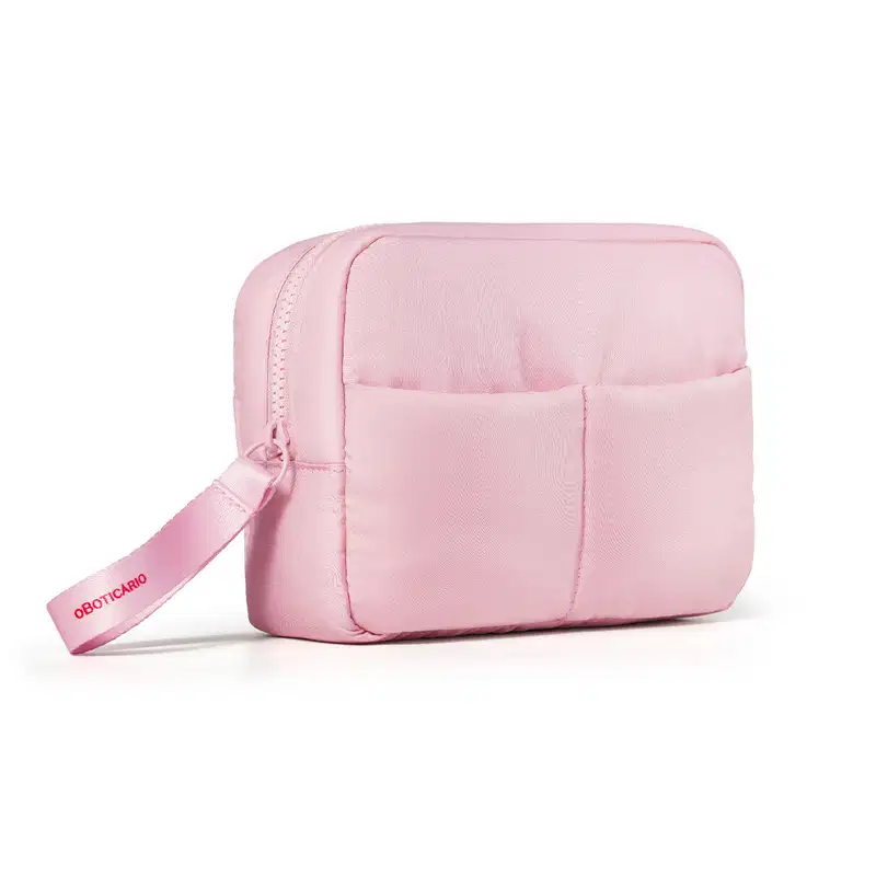 Imagem de uma nécessaire rosa clara com puxador. A bolsa tem vários compartimentos e um zíper na parte frontal. Há o nome da marca o Boticário em rosa escuro no puxador.