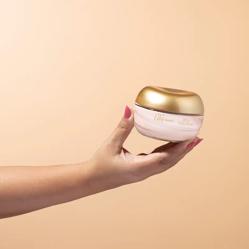 Imagem de uma mão feminina segurando um pote de vidro transparente com conteúdo branco e tampa dourada contendo creme acetinado corporal da marca L'eau de Lily Soleil em fundo bege vibrante.