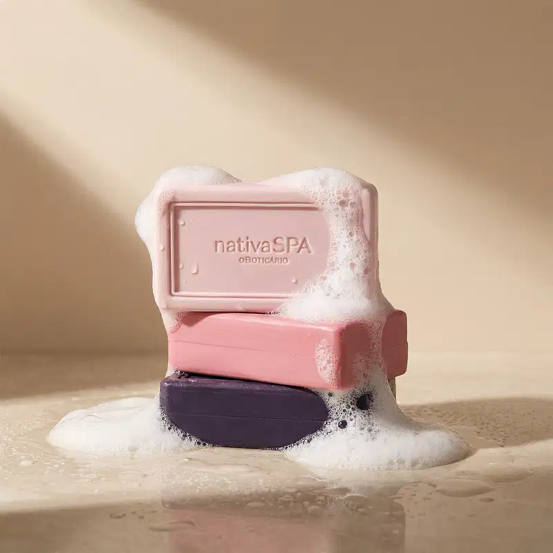 A imagem mostra três barras de sabonete em tons de rosa e roxo da marca Nativa SPA estão cheios de espuma branca, sobre superfície clara, sombreada e molhada.
