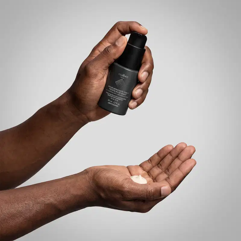 Mão segurando um frasco preto de item de perfumaria da marca Malbec sobre uma superfície clara, em um ambiente minimalista e monocromático.