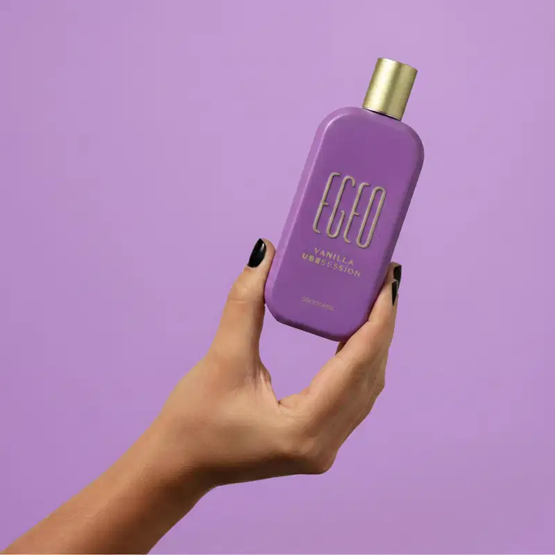 Frasco de item de perfumaria da marca Egeo em tom de roxo, com tampa dourada, segurado por mão feminina sobre fundo liso roxo.
