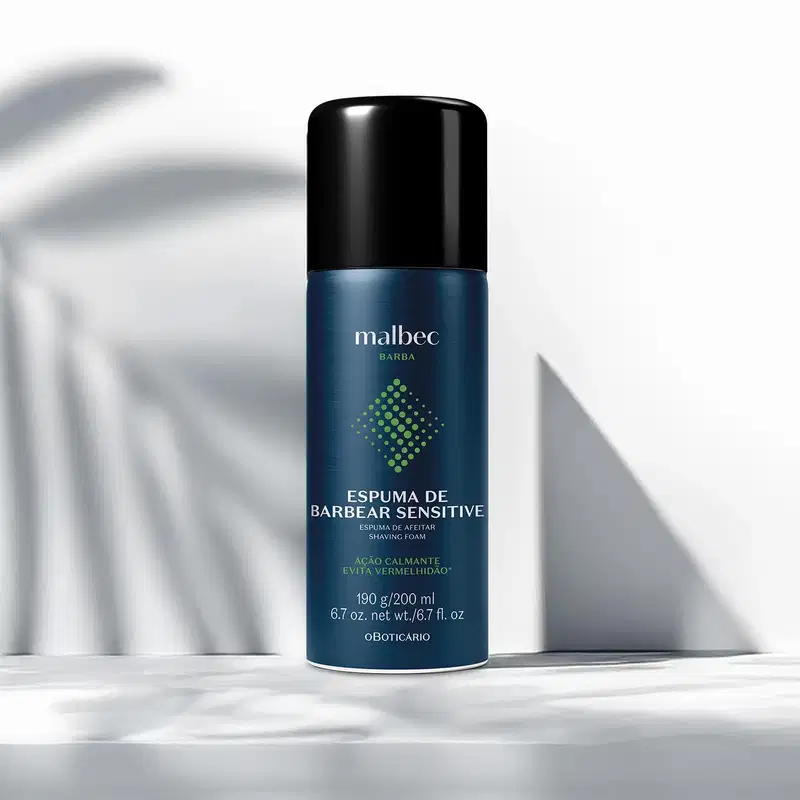 A embalagem verde metálica da espuma de barbear da marca Malbec está sobre superfície clara com sombras suaves, em ambiente minimalista. O produto possui rótulo em branco e dourado e tampa preta.