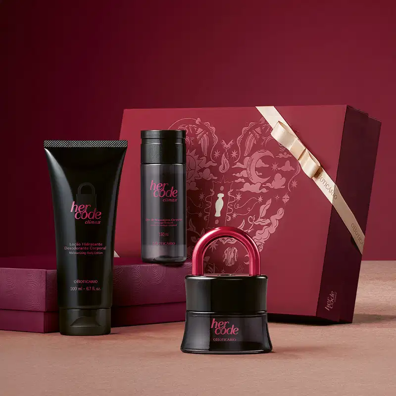 Caixa vermelha com ilustrações vista lateralmente e outra posicionada horizontalmente sobre superfície rosa clara e fundo vinho, com 3 itens de perfumaria da linha Her Code Clímax, sendo o frasco de desodorante colônia em primeiro plano.