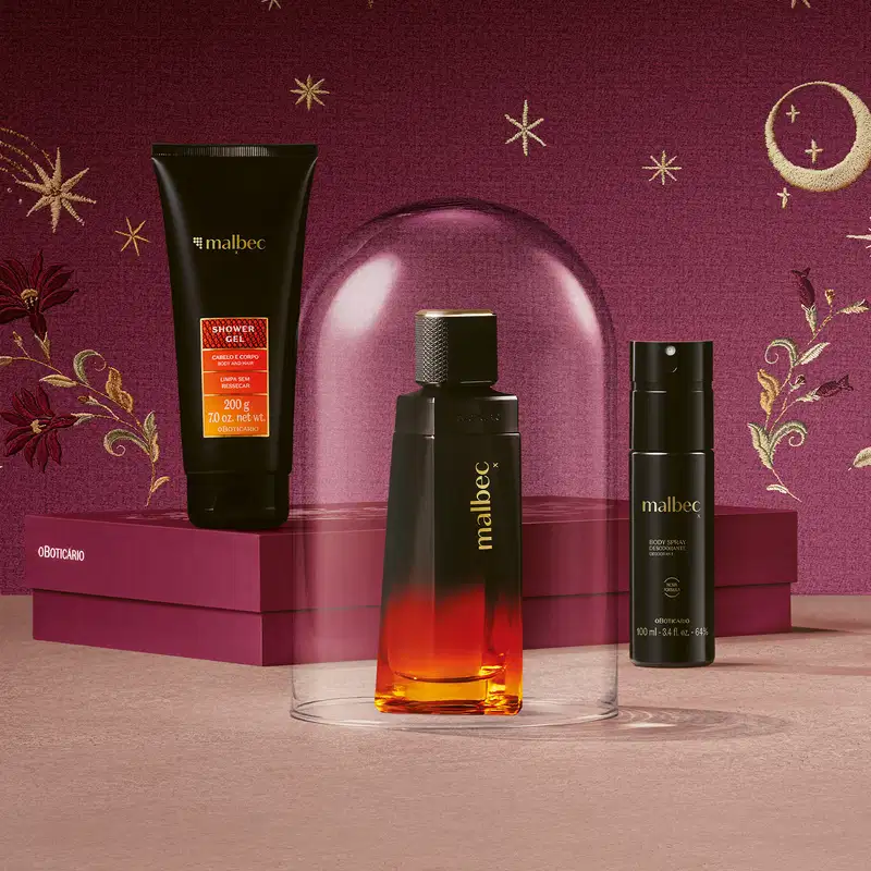 Produtos da linha Malbec X e caixa vinho posicionada horizontalmente sobre superfície rosa. Em primeiro plano, frasco de fragrância dentro de redoma transparente, e body spray ao lado e bisnaga de shower gel sobre a tampa da caixa em fundo rosa escuro.