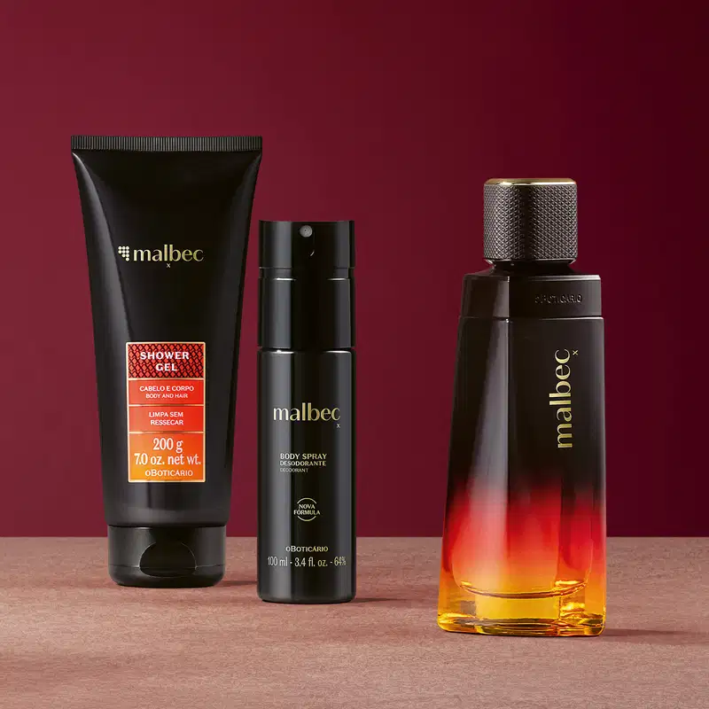 Três produtos de perfumaria da linha Malbec X em uma superfície rosa e fundo vinho, incluindo um frasco de item de perfumaria, um body spray e um gel de banho em embalagem preta e laranja.
