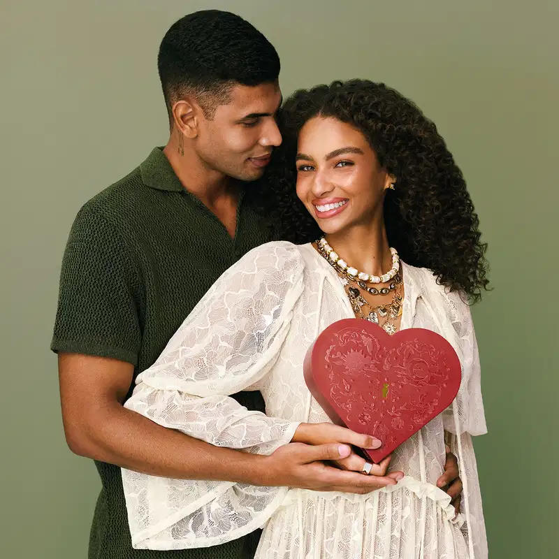 Casal negro jovem em fundo cinza, sendo que a mulher veste vestido branco de manga longa esvoaçante e segura uma caixa de presente em formato de coração vermelho enquanto o homem veste camiseta verde escuro e a abraça carinhosamente.