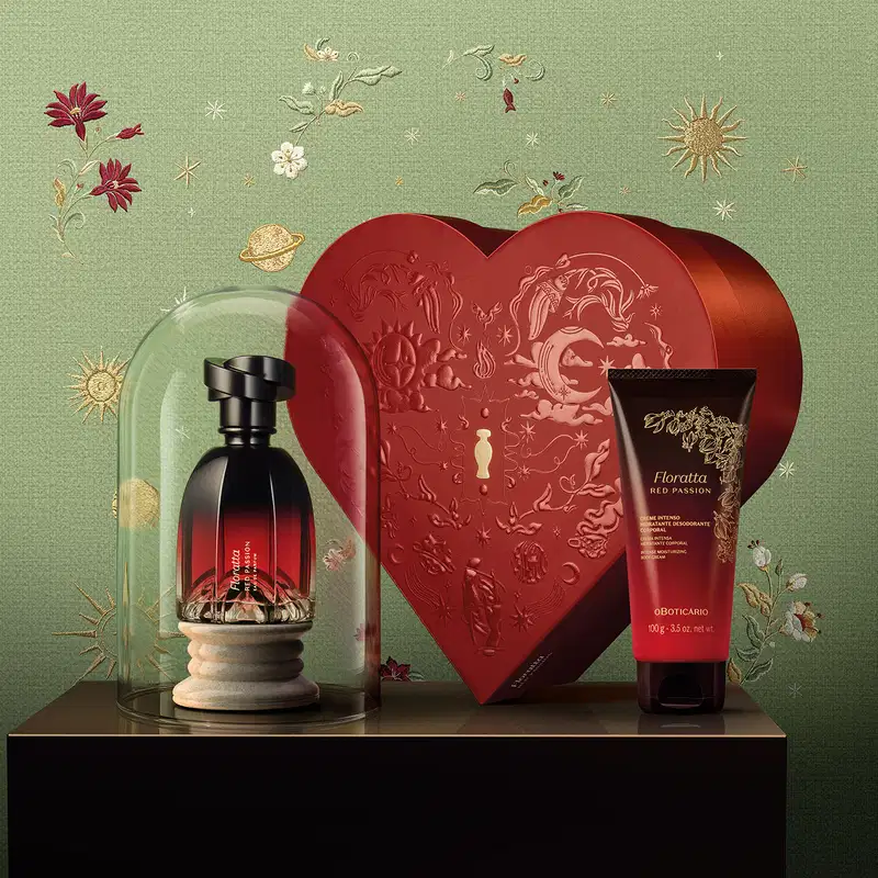 Cena de fundo verde com flores e estrelas douradas bordadas ao fundo. No primeiro plano, uma superfície marrom exibindo um frasco Eau de Parfum Floratta Red Passion dentro de uma redoma de vidro, uma bisnaga de creme corporal e caixa em formato de coração