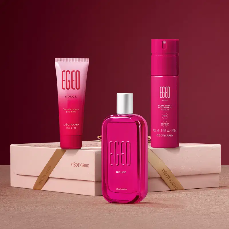 Caixa rosa clara posicionada horizontalmente sobre superfície rosa clara e fundo rosa escuro com três embalagens sobre ela, sendo em primeiro plano o frasco de desodorante colônia Egeo Dolce e ao fundo, a bisnaga e o body spray, todos na cor pink.