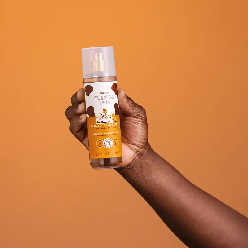Braço com mão segurando um frasco de body splash Cuide-se Bem Deleite Caramelizado em fundo laranja. O frasco possui design moderno, com rótulo branco e marrom e informações em letras brancas e pretas.