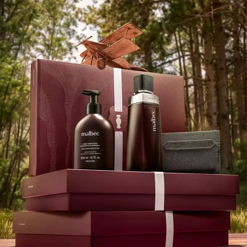 Caixa de presente com produtos de beleza da marca Malbec, em ambiente externo com vegetação ao fundo. Embalagem em tons de vermelho e preto, com desodorante, loção hidratante e perfume, sugerindo um conjunto de cuidados pessoais.