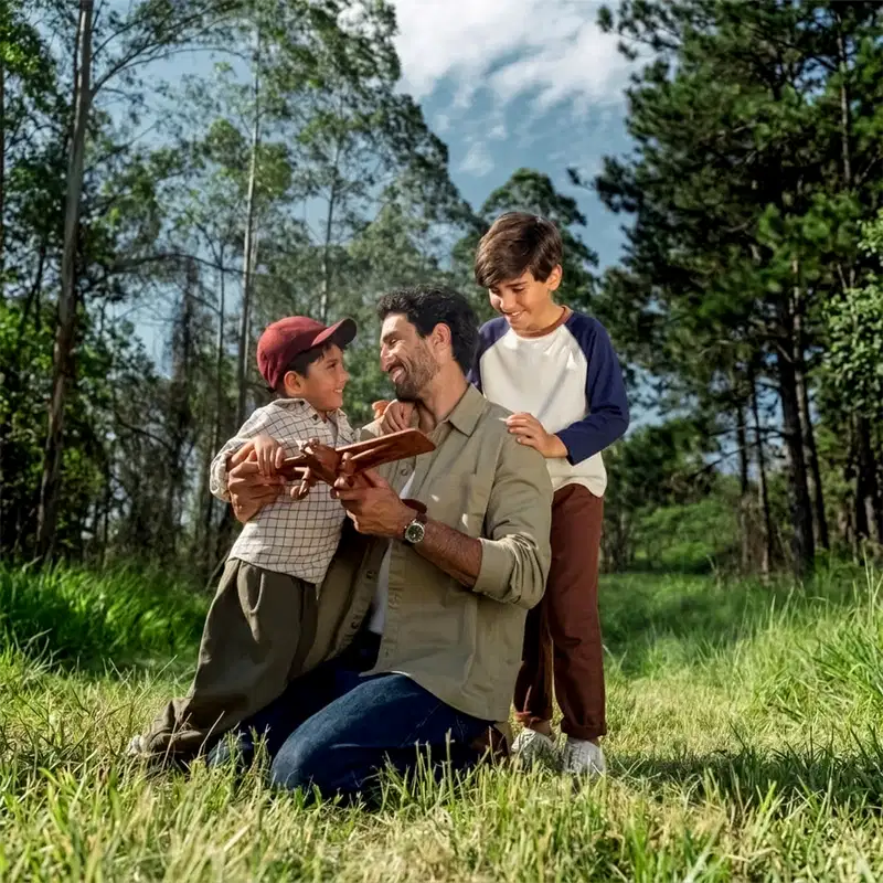 Um homem de camisa bege e calça marrom segura um livro e conversa com dois meninos em um ambiente natural com árvores e vegetação ao redor.