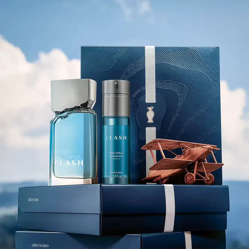 Caixa de presente azul-marinho com logotipo da linha Clash, contendo item de perfumaria e embalagem complementar, sobre fundo de céu com nuvens e avião de madeira.