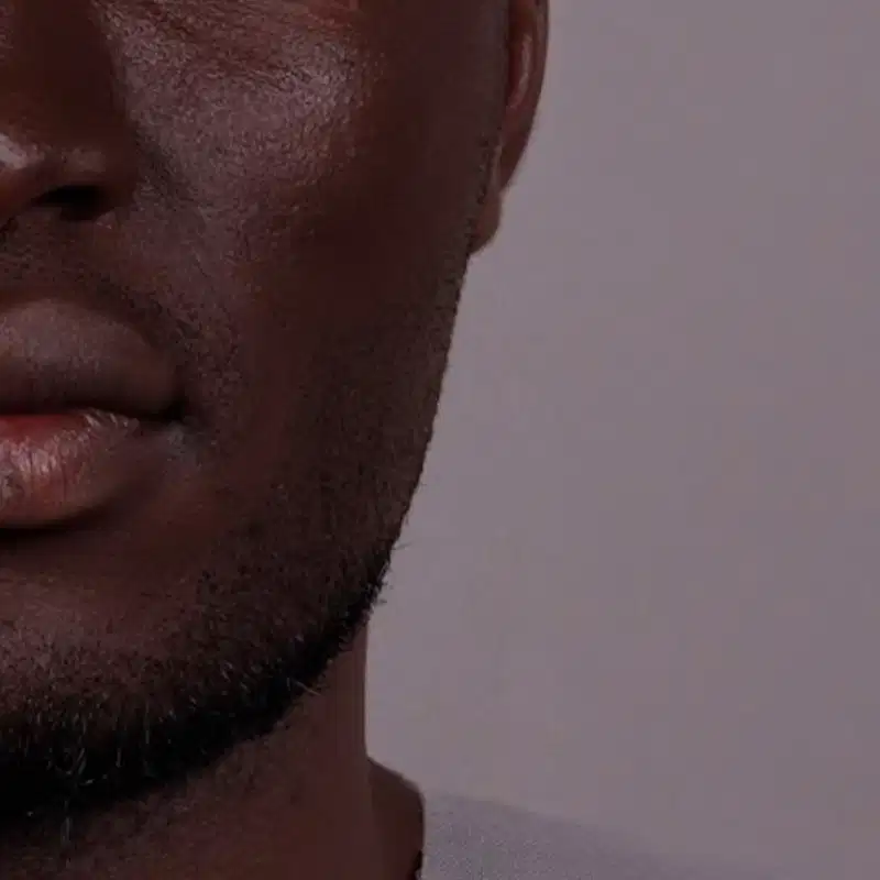 Imagem de close-up do quarto inferior esquerdo de um rosto masculino negro com barba rente e camiseta de gola redonda branca, em um fundo cinza claro.