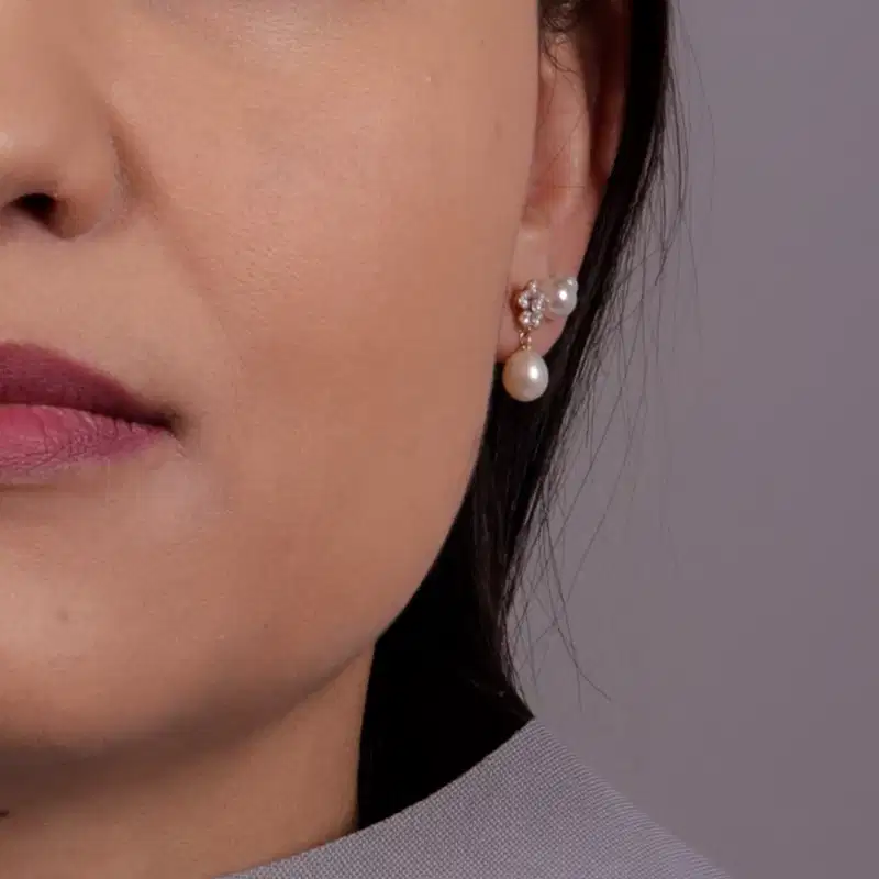 Imagem de close-up do quarto inferior esquerdo de um rosto feminino branco com cabelo longo, castanho e liso com brinco pequeno de pedrarias e pendente de pérola na orelha em fundo cinza claro.