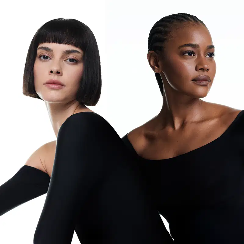 Duas modelos vestindo roupas pretas, sendo uma mulher negra com cabelo trançado preso e outra branca com cabelo curto preto e franja em pose empoderada em fundo branco.