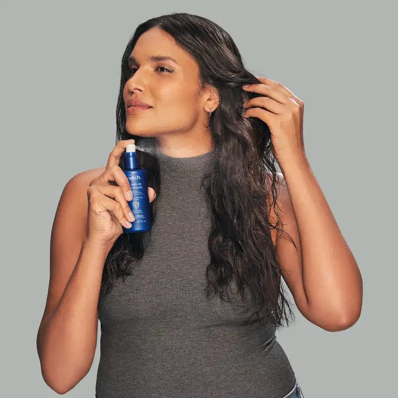 Mulher segurando um item de cabelos azul em frente ao corpo, vestindo uma blusa cinza em um fundo cinza claro. Cabelos longos e ondulados. Ambiente neutro e minimalista que destaca o produto.