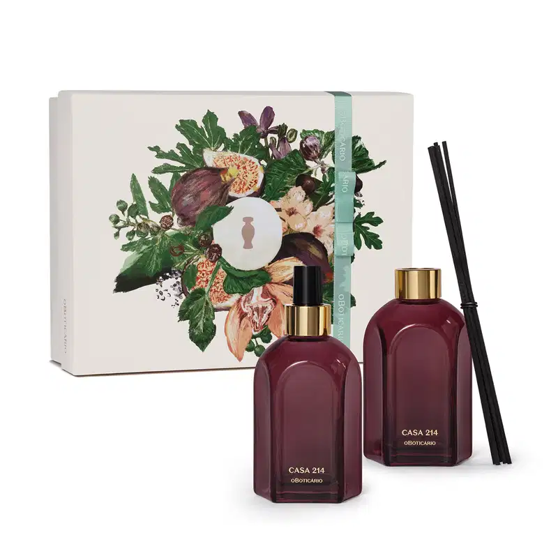 Caixa branca com ilustração floral e dois vidros retangulares na cor roxa, com rótulo em dourado. Um deles é o aromatizador de ambientes com válvula spray preta e o outro é um difusor com tampa dourada que acompanha varetas de madeira pretas.