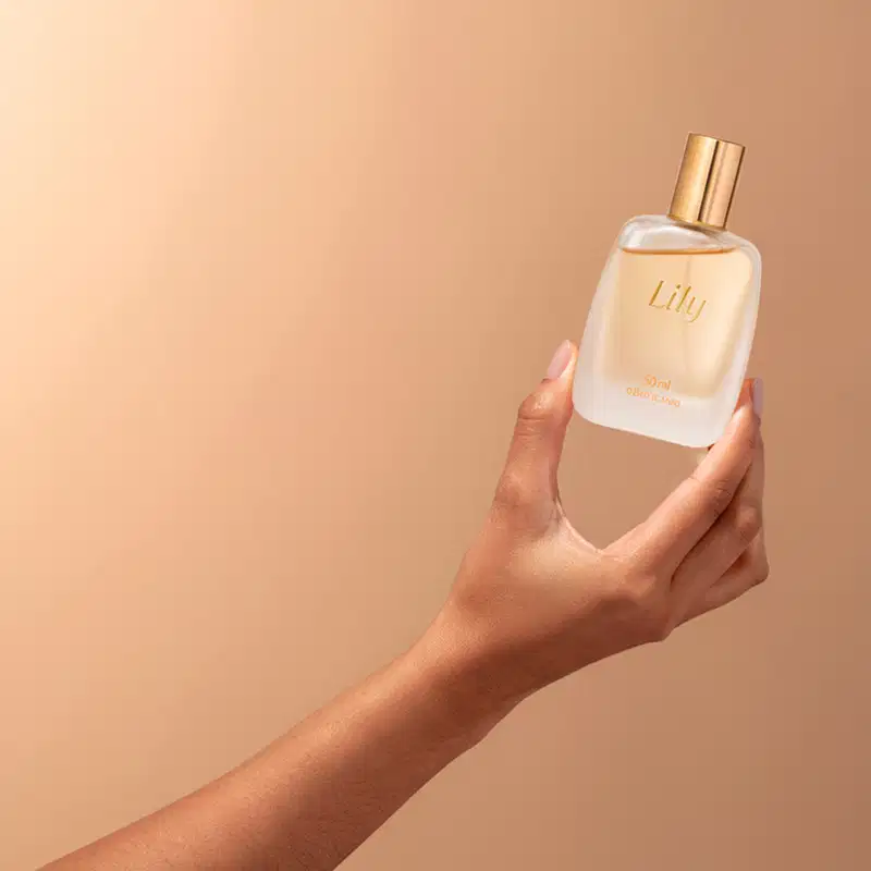 Imagem de um frasco de vidro da fragrância Lily em embalagem transparente com tampa e letras douradas, sendo segurado por uma mão feminina com fundo bege.