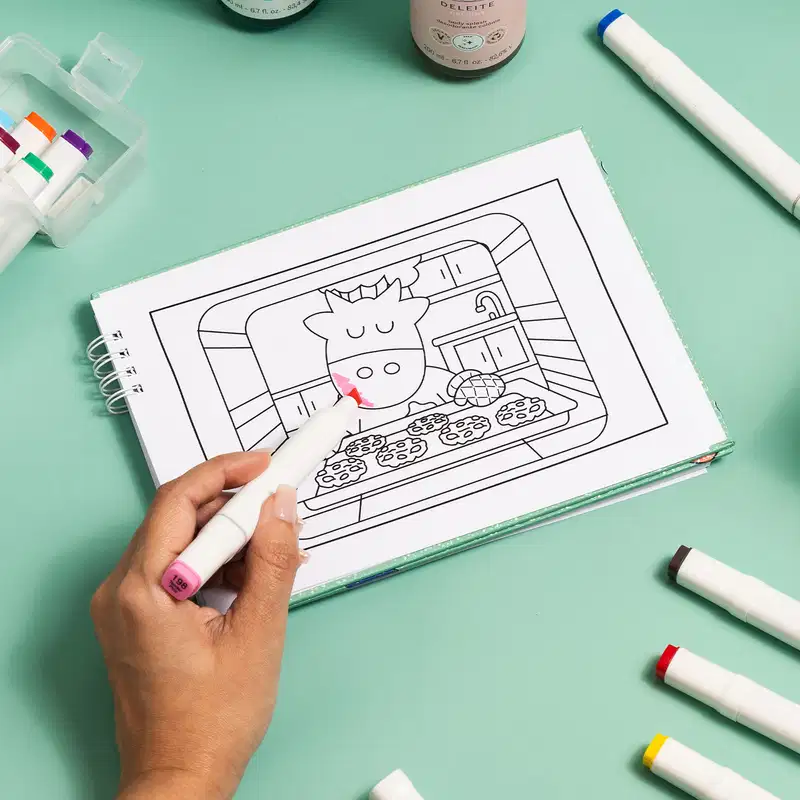 Imagem de uma superfície verde clara com diversas canetinhas coloriadas ao redor de um livro de colorir com a imagem de uma vaquinha cozinhando cookies. Uma mão com uma canetinha rosa pinta o focinho da vaca enquanto o resto da imagem segue preto e branco