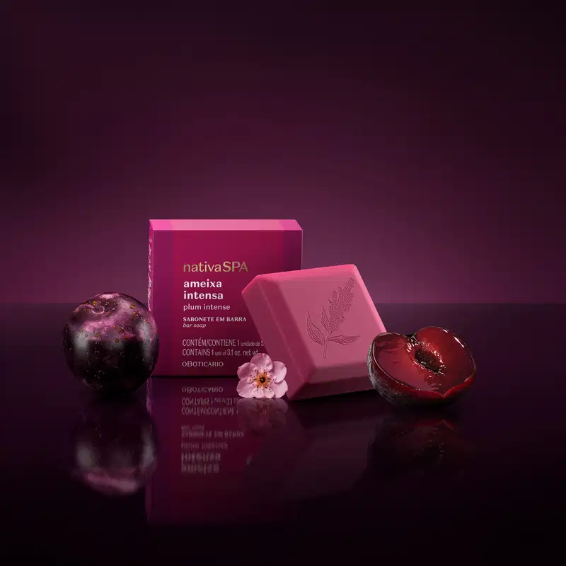 Embalagem de sabonete em barra da marca Nativa SPA, em tom de rosa intenso, com uma flor rosa, cercada por ameixas roxas em fundo escuro e reflexivo.