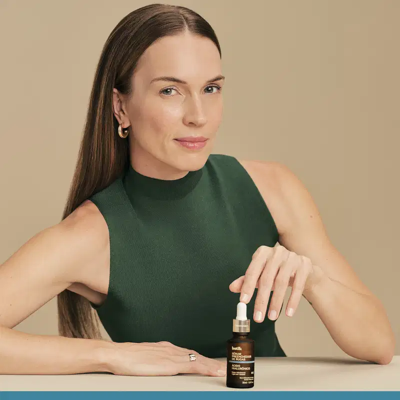 Uma mulher com cabelos longos e escuros, vestindo uma blusa verde escura, segurando um frasco de vidro com um item de perfumaria da marca Botik em um fundo bege.