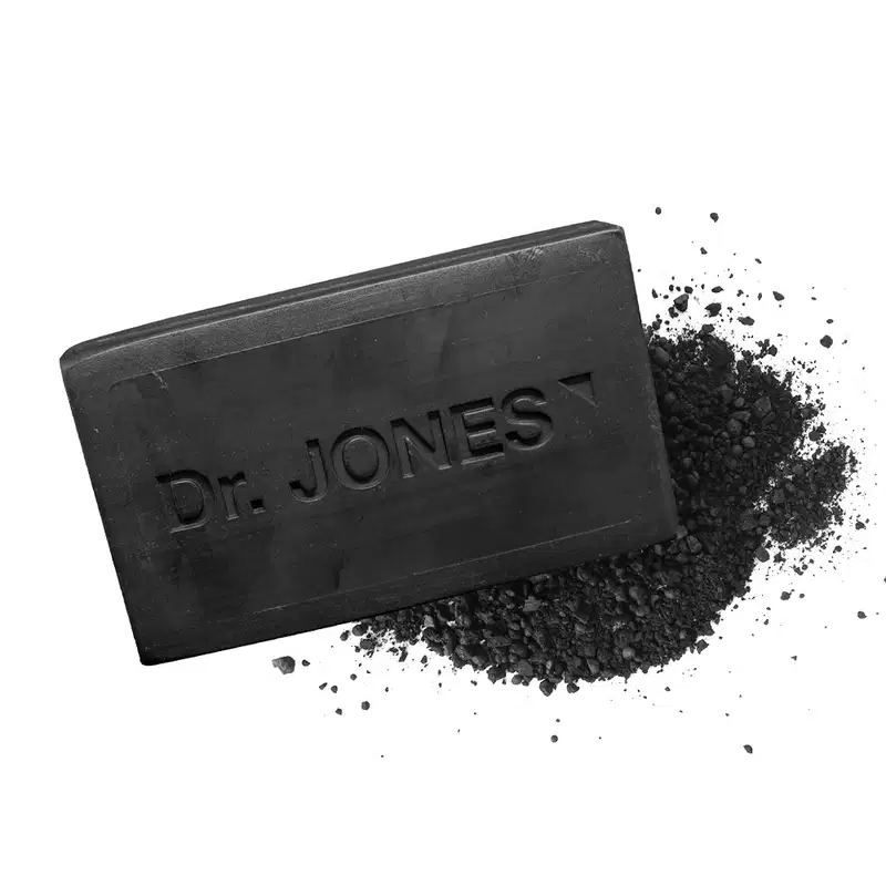 A imagem mostra o sabonete em barra na cor preta com a inscrição "Dr. Jones" em relevo. Ao lado há uma quantidade significativa de carvão espalhado em forma de grânulos.