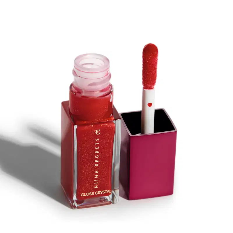 Frasco de vidro retangular transparente contendo um o gloss brilhante vermelho com partículas cintilantes. O frasco está aberto e ao lado está o aplicador labial na mesma cor vermelha cintilante acoplado à tampa compacta de cor rosa escuro.