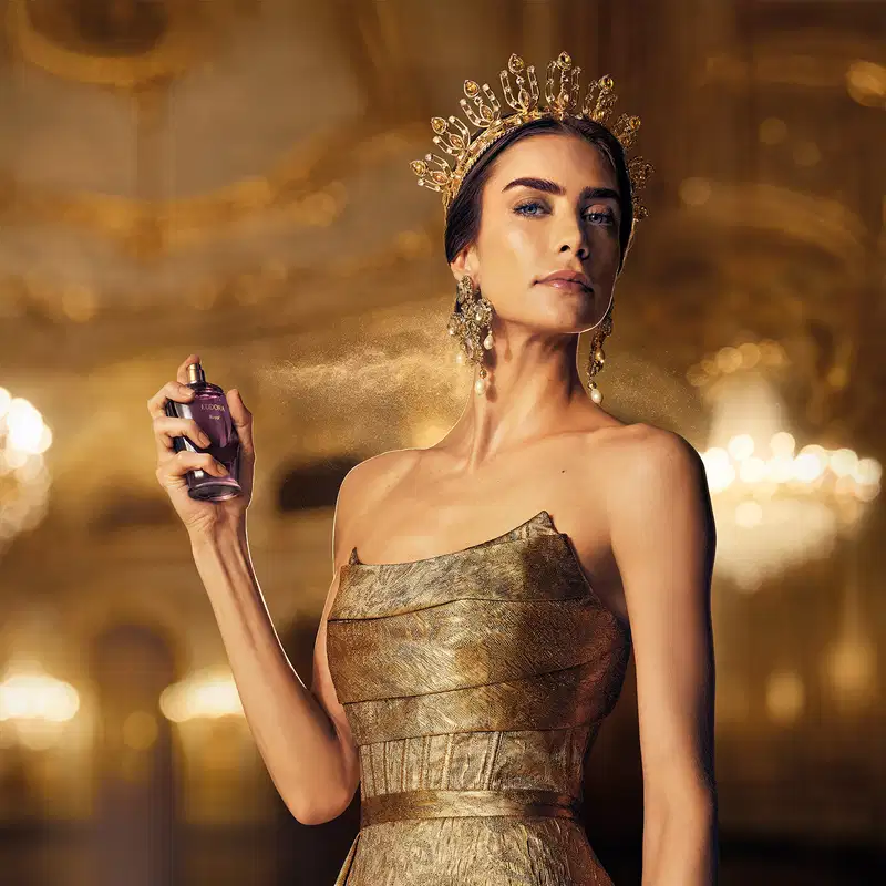 Ambiente de luxo e glamour com iluminação dourada, retratando uma mulher usando uma coroa ornamentada e segurando um frasco de Eudora Royal, em um cenário que sugere uma ocasião de realeza.