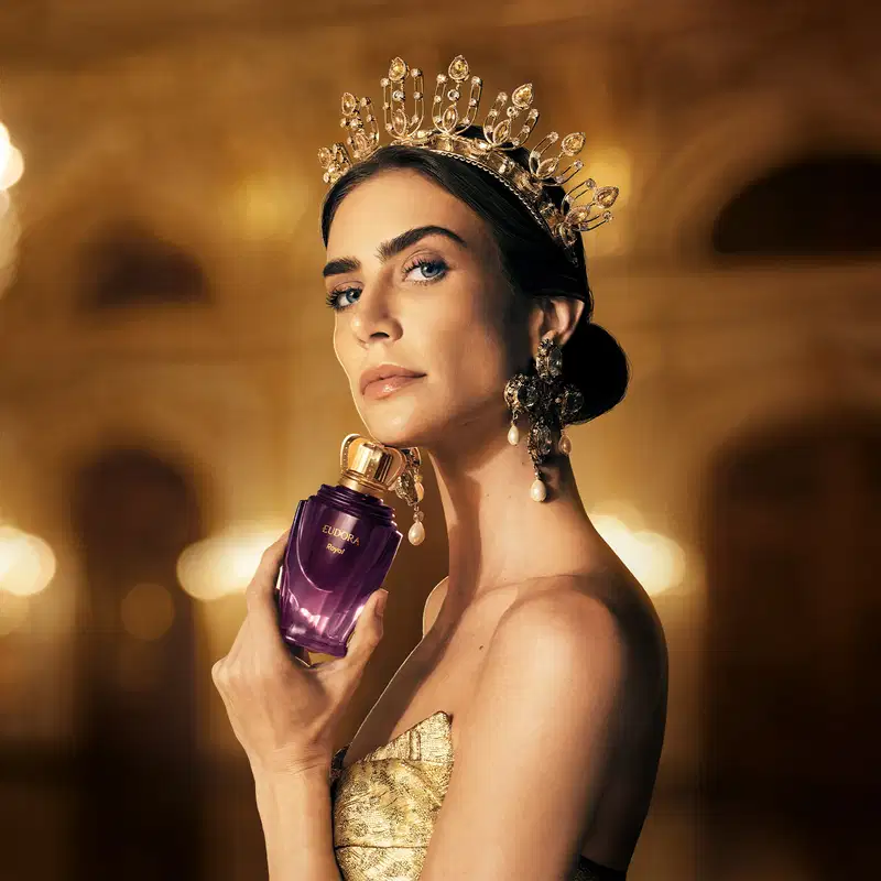 A modelo usa um elegante vestido dourado e uma coroa cintilante, segurando um frasco de Eudora Royal em um ambiente luxuoso e dourado, sugerindo uma ocasião da realeza.