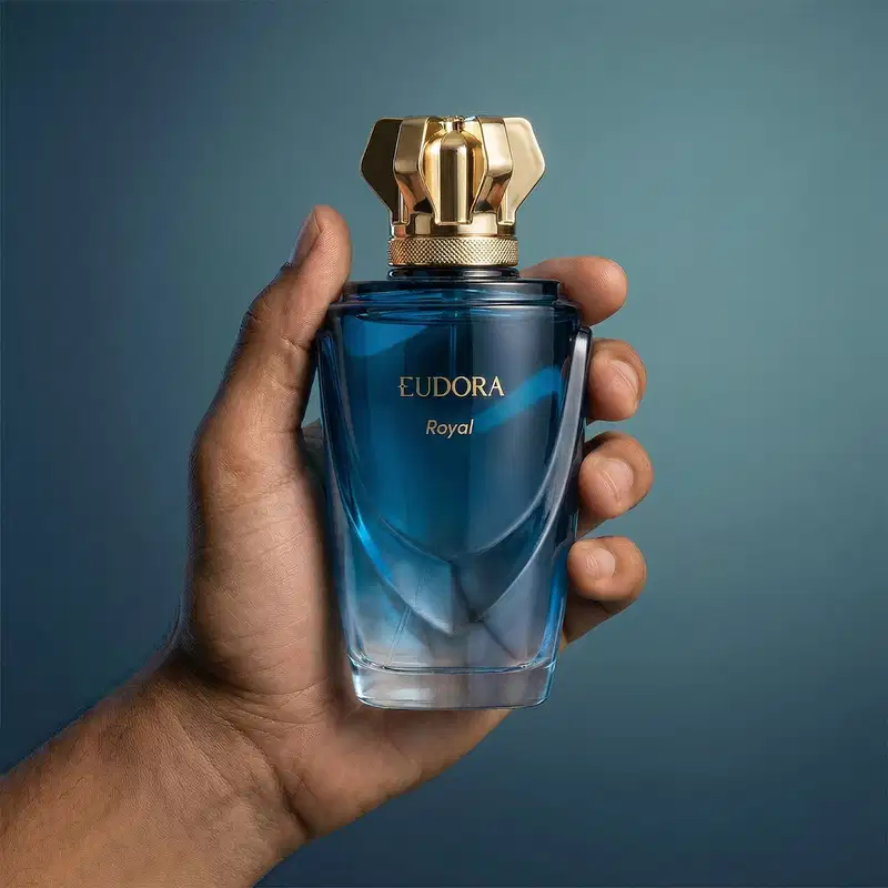 Frasco azul da marca Eudora, com tampa dourada em formato geométrico. Rótulo com o nome "Eudora Royal" em letras douradas. Produto segurado em mãos masculinas contra fundo azul escuro.
