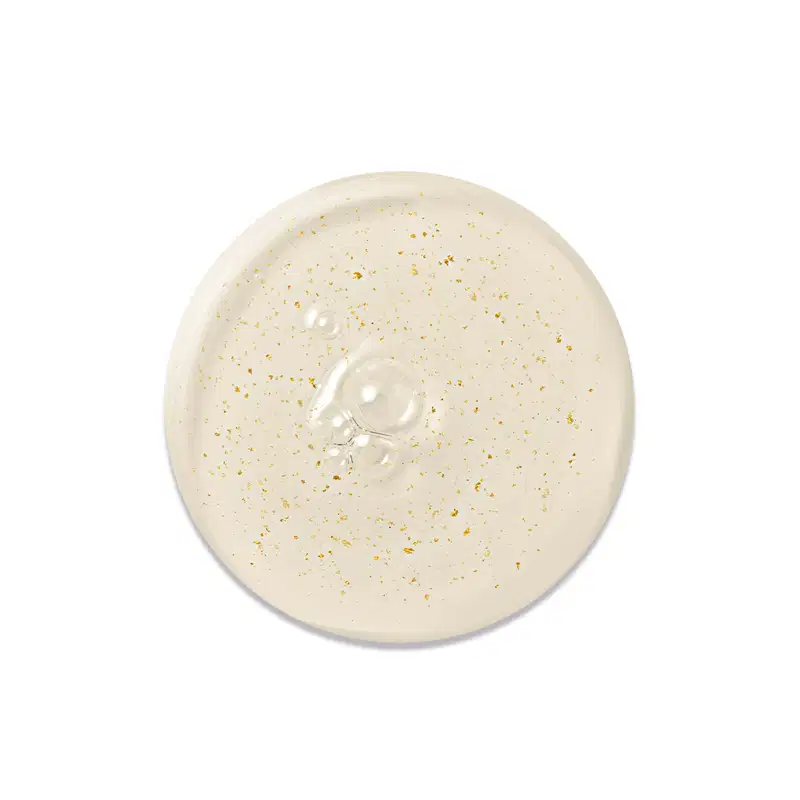 Textura do Primer. Gota bege claro com partículas douradas espalhadas por toda a superfície. Formato circular com diâmetro aproximado de 6 cm. Superfície levemente côncava no centro, onde se concentram algumas gotas de um produto líquido translúcido.