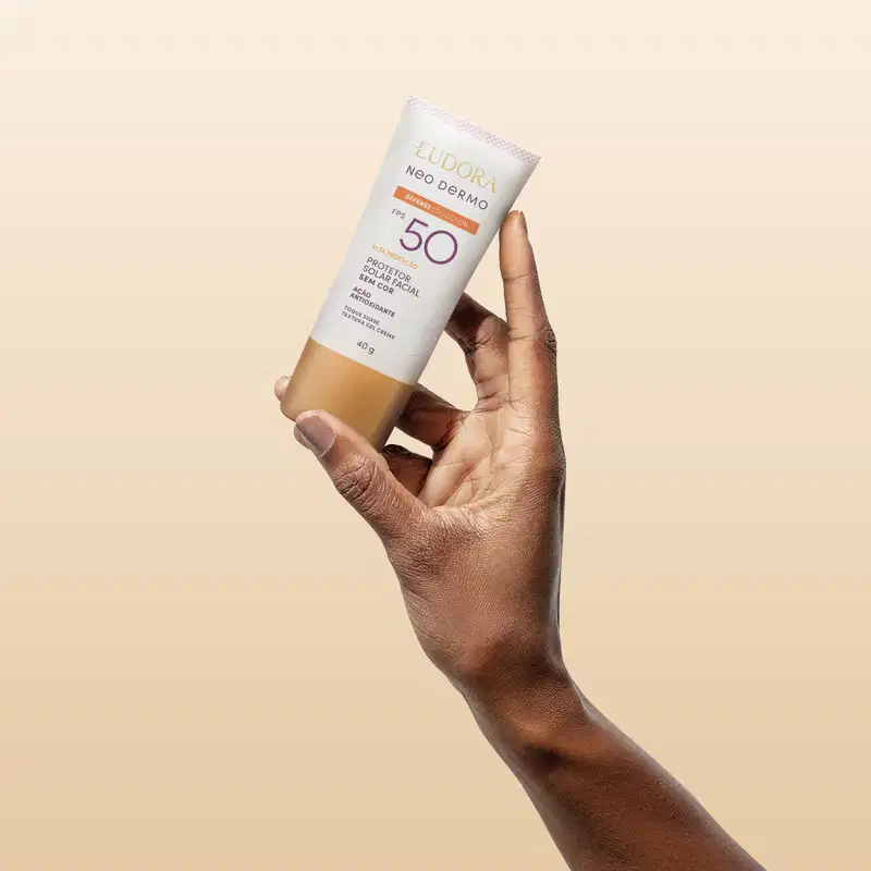 O produto mostrado está sendo segurado por uma mão, é um protetor solar Neo Dermo com FPS 50, em um tubo branco com detalhes roxo e laranja.