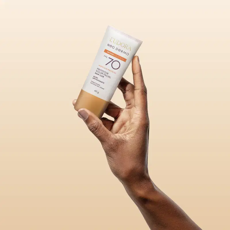 O produto mostrado está sendo segurado por uma mão, é um protetor solar Neo Dermo com FPS 50, em um tubo branco com detalhes roxo e laranja.