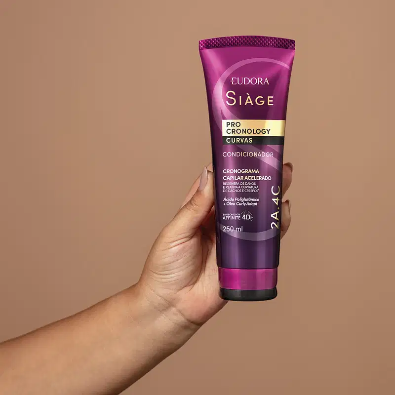 O produto mostrado na imagem está sendo segurado na mão. É um condicionador da linha Siàge Pro Cronology Curvas, da marca Eudora. O condicionador está em uma embalagem de tubo plástico de cor roxa e rosa, com detalhes texturizados.