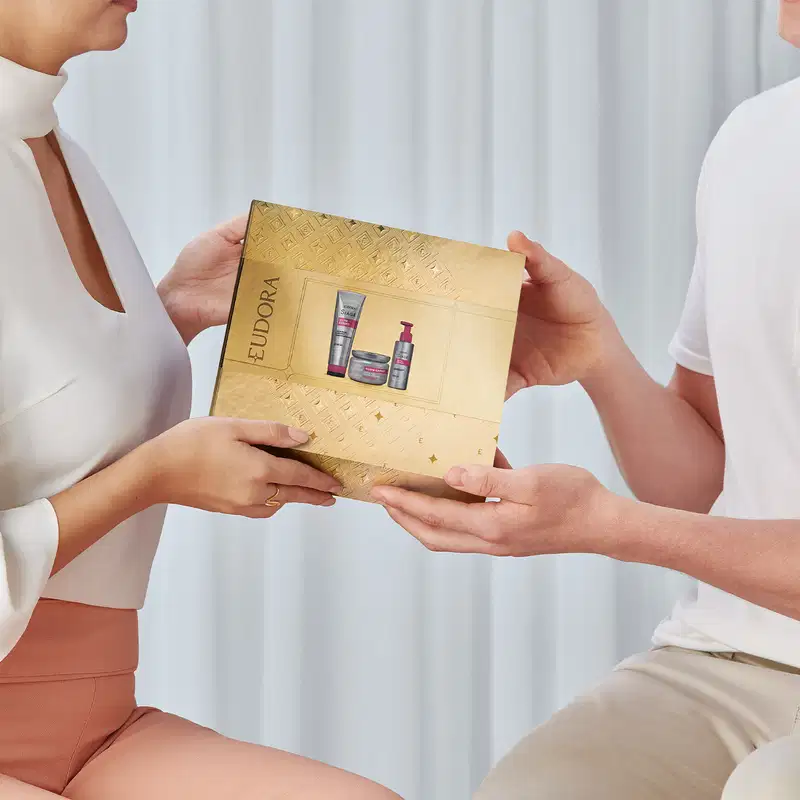A imagem mostra um homem e uma mulher segurando uma caixa dourada, como se estivesse entregando um presente.