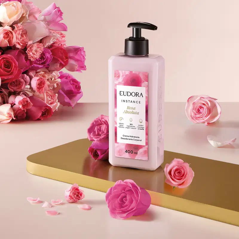 O frasco possui um design moderno, com uma bomba dispensadora preta. A embalagem tem uma coloração rosa clara, com detalhes em dourado. Ao redor do frasco, há diversos arranjos de rosas em tons de rosa, complementando a apresentação.