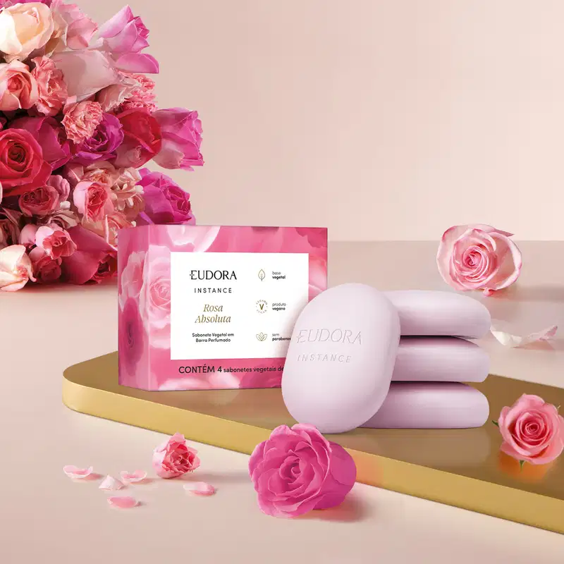 Caixa de sabonetes vegetais da linha Instance Rosa Absoluta. A caixa rosa contém 4 sabonetes vegetais perfumados. A imagem também mostra algumas rosas frescas em tons de rosa, criando um cenário delicado e floral.