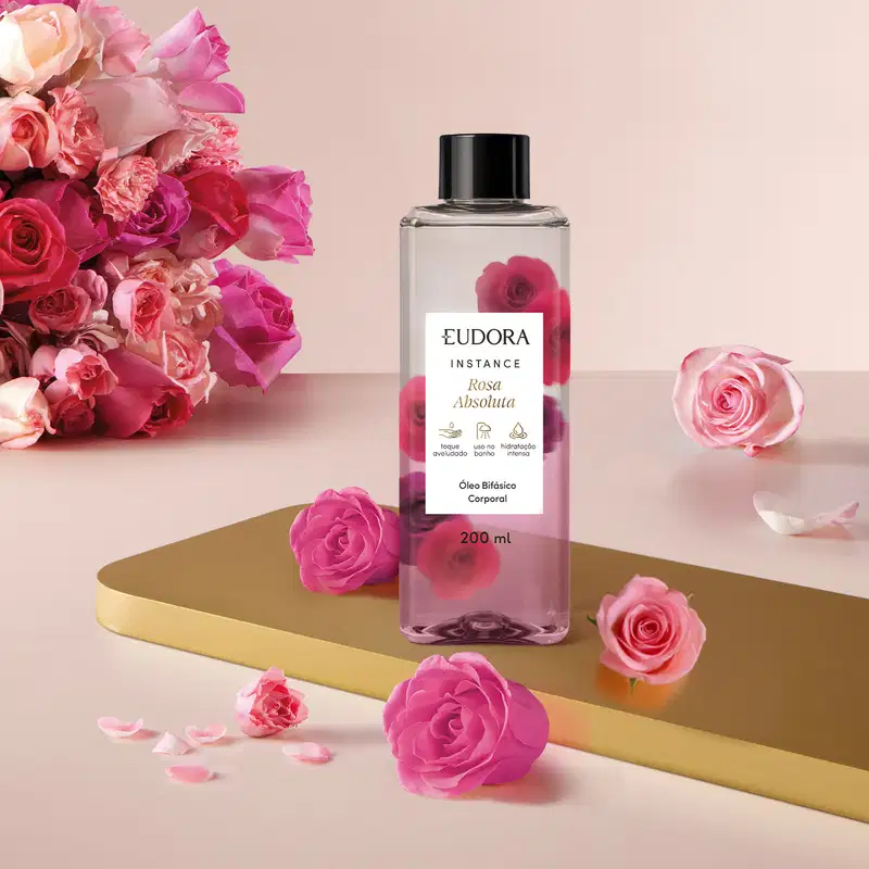 O frasco do Óleo Bifásico Instance Rosa Absoluta é transparente, com um rótulo preto e branco, e contém um líquido claro. O produto está posicionado em um ambiente com rosas rosa, criando uma composição visual harmoniosa.